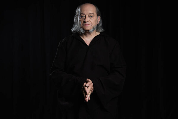 medieval monk in dark cassock standing with folded hands and looking at camera isolated on black