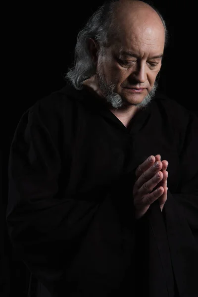 senior medieval monk in dark robe praying isolated on black