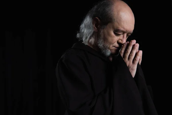 senior bearded monk in robe praying with closed eyes isolated on black