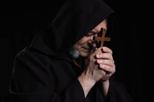 senior monk in hooded cassock praying with crucifix near face isolated on black