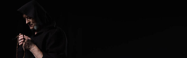 монах в темном сутане в капюшоне молится с четками изолированы на черном, баннер
