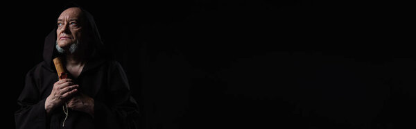 monk holding medieval manuscript and looking away isolated on black, banner