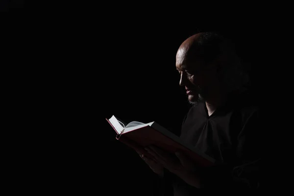 mysterious monk in robe reading bible in darkness isolated on black