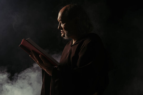 side view of senior priest in cassock reading holy bible on black with smoke