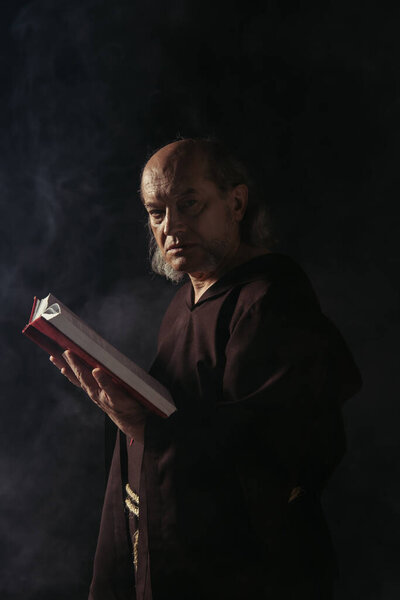 строгий старший монах смотрит в камеру, держа Библию на черном от дыма