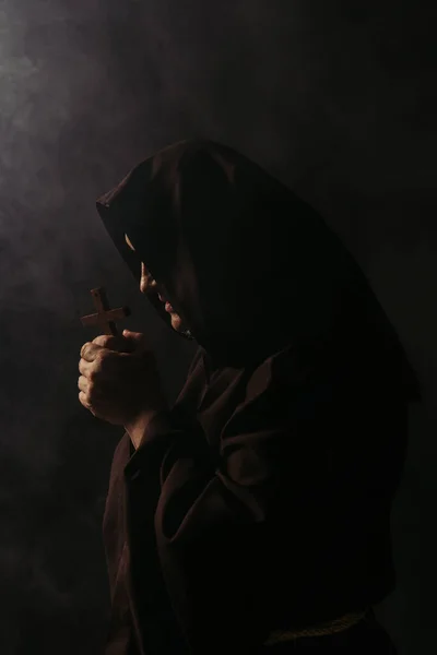 mysterious medieval monk in hooded robe praying with crucifix on black background