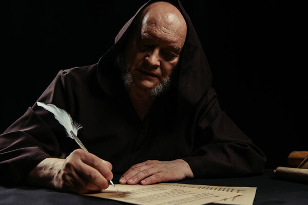 priest in dark hooded cassock writing manuscript at night isolated on black