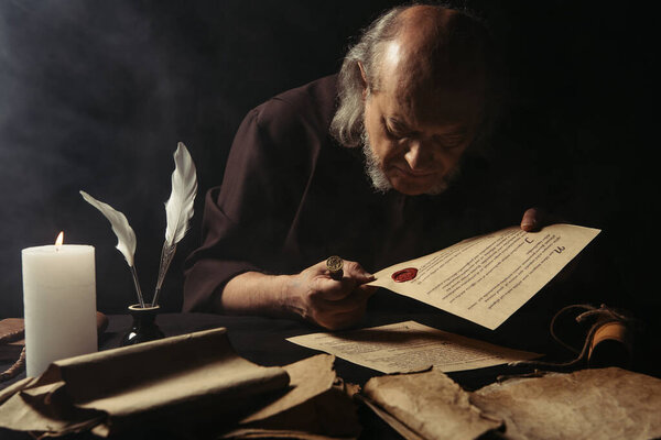 senior monk looking at chronicle stamped with wax seal near parchments and burning candle on dark background