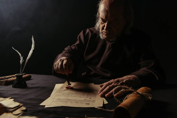 ancient monk stamping manuscript with wax seal near rolled parchment isolated on black