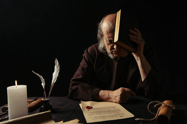 senior monk obscuring face with bible near chronicle with wax seal isolated on black