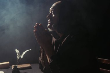 Ortaçağ keşişi mürekkep tenceresinin yanında dua ediyor ve siyah arkaplanda dumanla el yazması yazıyor.