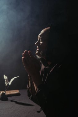 Tespih boncuklu kıdemli rahip mürekkep küpünün yanında tüy kalemleriyle dumanla dua ediyor.
