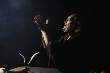 Ortaçağ keşişinin siyah arka planda oklu mürekkep küpünün yanında dua edişinin yan görüntüsü