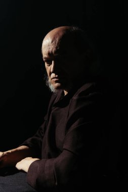 Ciddi ve düşünceli bir rahip oturup siyah kameraya bakıyor.
