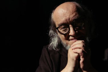 Gözlüklü gülümseyen filozof ve sıkıca sarılmış elleriyle siyah kameraya bakıyor.