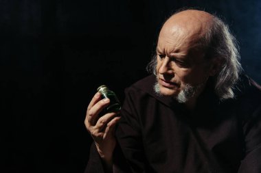 Düşünceli simyacı, siyah üzerine sihirli malzeme konmuş kavanoza bakıyor.