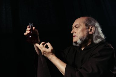Sakallı simyacı elinde iksirle siyah bir şişe tutuyor.