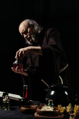 alchemist in mantle holding flask with liquid near pot and dried herbs isolated on black