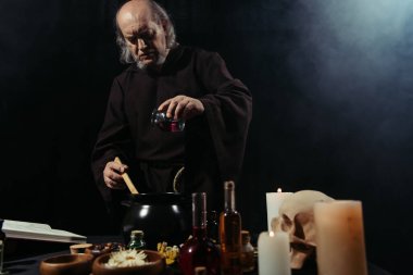 esrarengiz simyacı ot içerken siyah arkaplanda dumanla yemek pişiriyor.