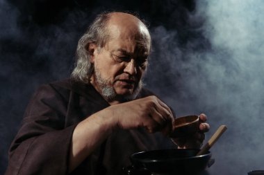 Sakallı simyacı ot içerken siyah arkaplanda dumanla iksir hazırlıyor.