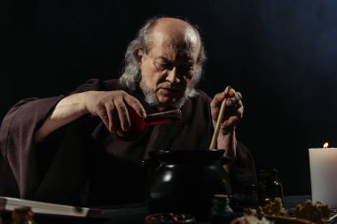 Gizemli simyacı sihirli iksir hazırlıyor ve siyah tencereye sıvı döküyor.