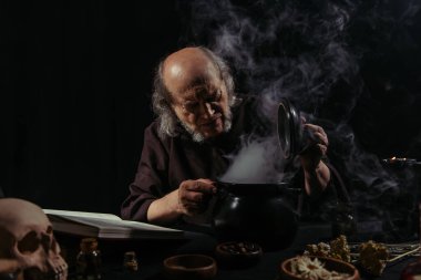 medieval alchemist near boiling pot and magic cookbook on black background clipart