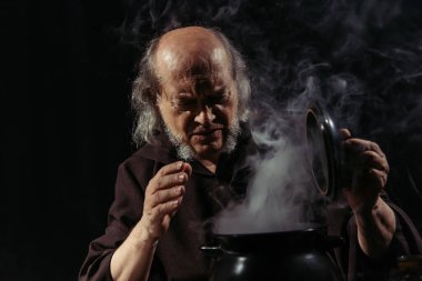 senior alchemist frowning near steaming pot isolated on black clipart