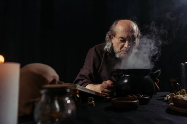 senior alchemist near boiling and steaming pot preparing potion at night isolated on black clipart