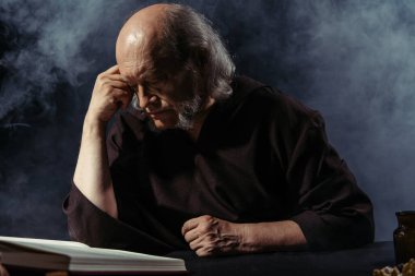 Kıdemli simyacı gece sihirli yemek kitabı okuyor. Siyah arka planda dumanlı.