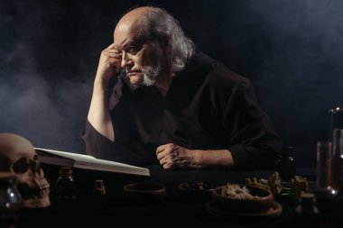 Ortaçağ simyacısı sihirli yemek kitabı ve kara arka planda kurutulmuş otları dumanla düşünüyor.