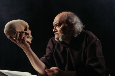 senior philosopher in dark robe looking at human skull isolated on black clipart