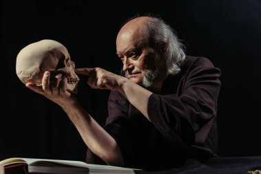 senior medieval philosopher pointing at human skull isolated on black clipart