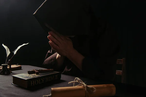 priest obscuring face with praying hands near crucifix on holy bible isolated on black