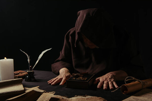 senior abbot in hood sitting near bible and medieval manuscripts isolated on black