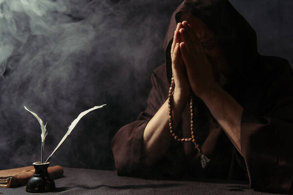 monk with rosary obscuring face with praying hands near inkpot and manuscript on black with smoke