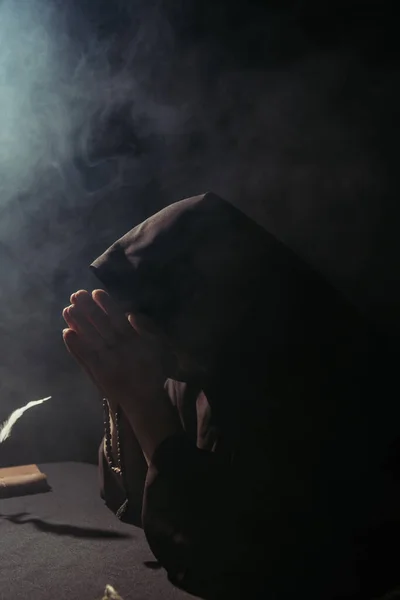 monk with obscured face praying with rosary at night on black with smoke