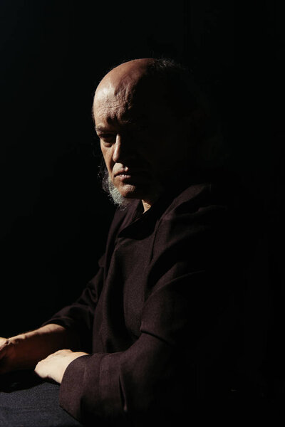 серьезный и вдумчивый священник сидит и смотрит на камеру, изолированную от черного