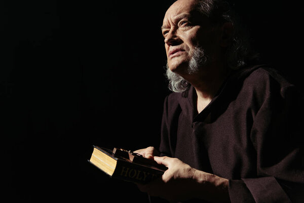 bearded monk in cassock holding bible and crucifix while thinking isolated on black