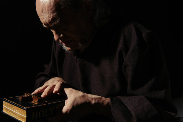 monk in dark robe holding bible and crucifix isolated on black