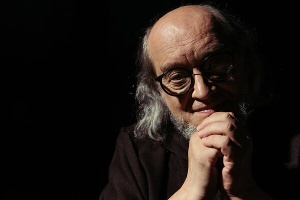 улыбающийся философ в очках и с сжатыми руками, смотрящий на камеру, изолированную на черном