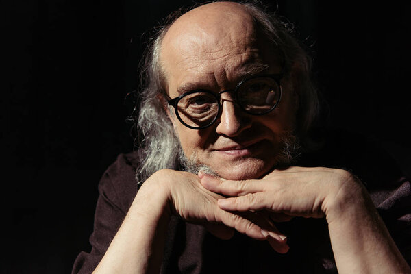 позитивный средневековый философ в очках, держась за руки возле подбородка и улыбаясь в камеру, изолированную на черном