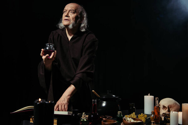 thoughtful alchemist holding jar with liquid near magic cookbook on black background 