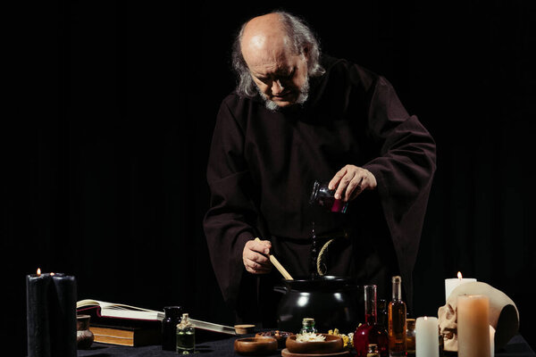 medieval alchemist preparing magic potion while adding ingredient into pot isolated on black