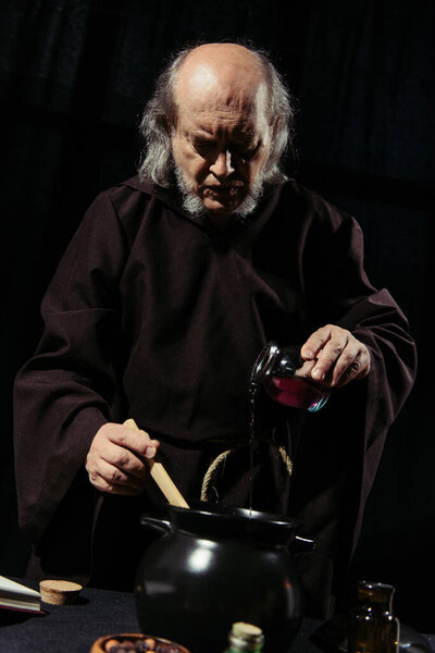 alchemist in dark robe pouring liquid into pot while cooking at night isolated on black