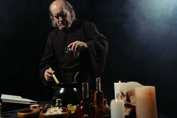 mysterious alchemist adding ingredient into pot while cooking in dark on black background with smoke