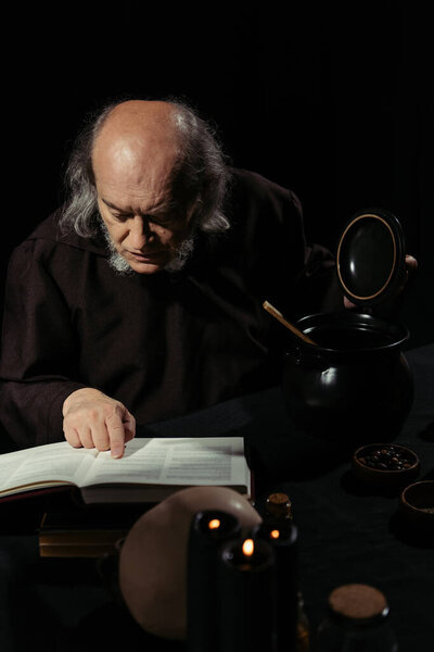 senior alchemist reading magic cookbook near pot isolated on black
