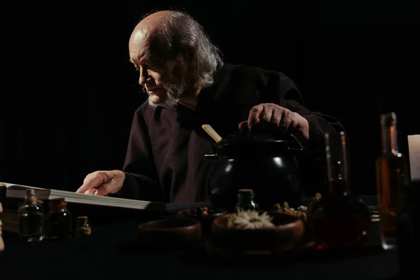 中世纪的巫师在锅和用黑色隔离的配料附近阅读魔法食谱 — 图库照片