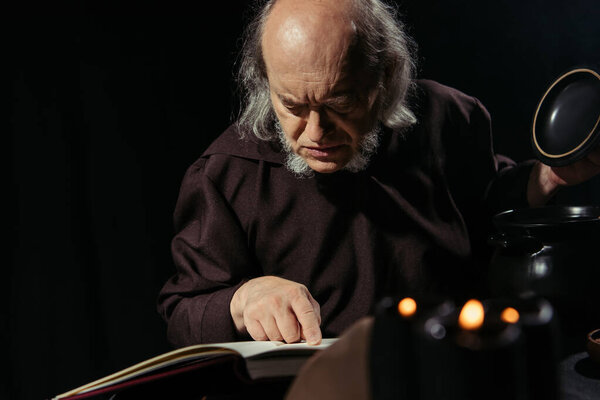 mysterious alchemic reading magic recipe in cookbook near pot isolated on black