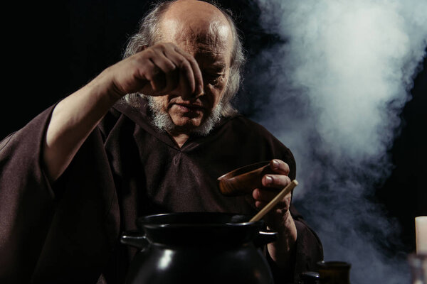 senior magician adding ingredient from bowl into pot on black background with smoke