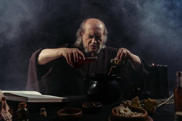 senior magician pouring liquid into pot while preparing potion on black background with smoke